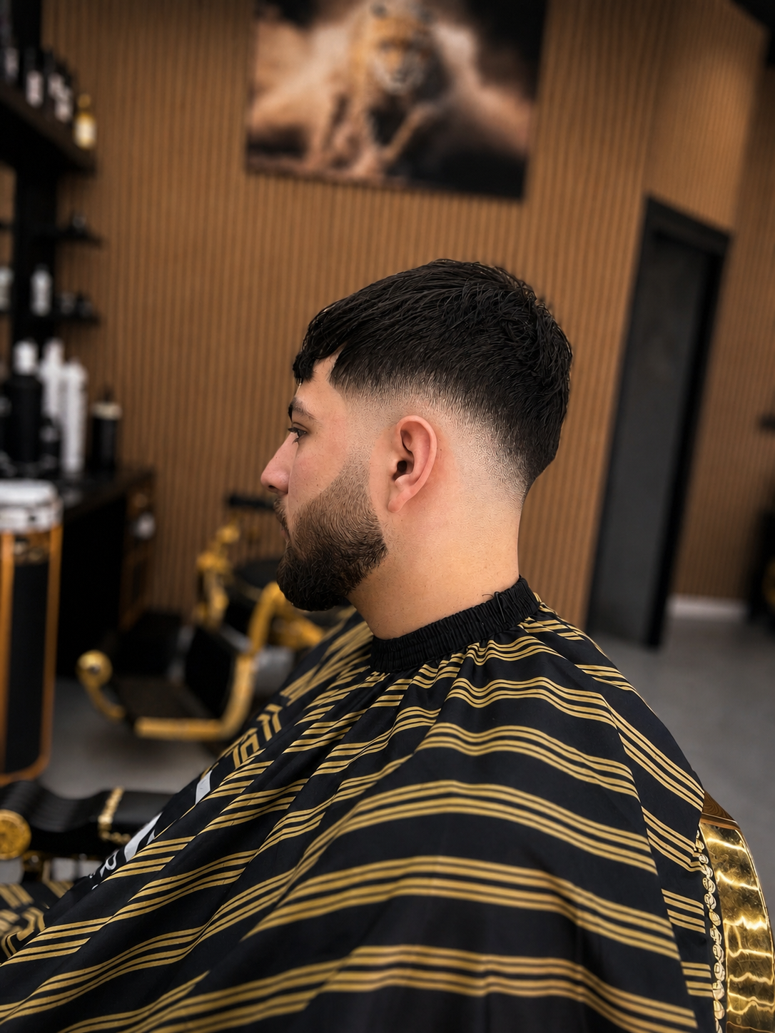 Klant van opzij met een korte fade en verzorgde baard, in een zwart-goud gestreepte kapmantel